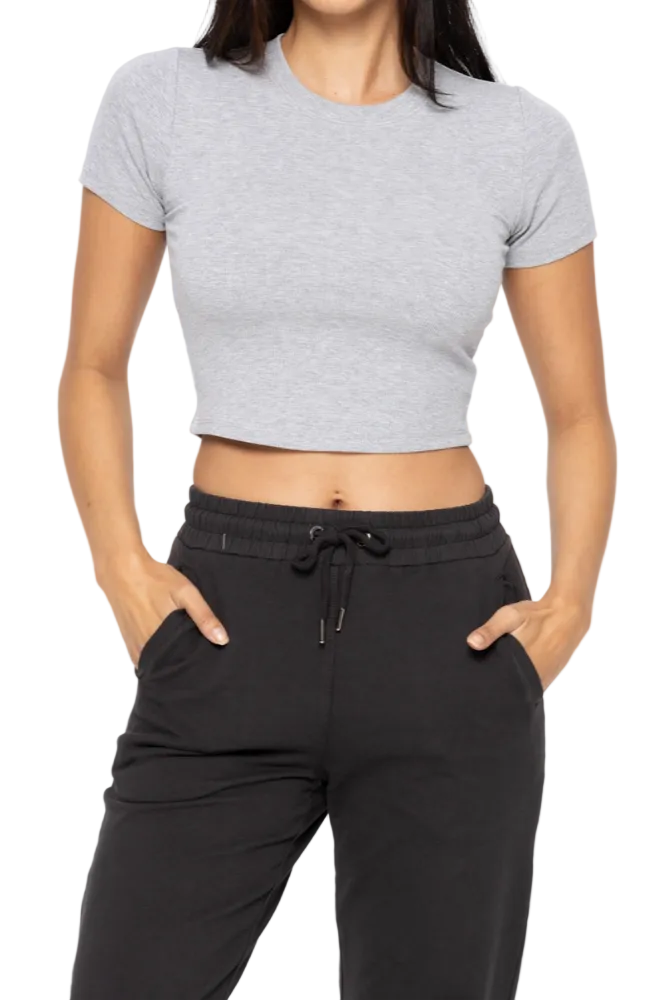 A woman wearing a cropped grey athleisure baby tee with short sleeves and a rounded neckline, paired with black leggings. She has a sleek, casual style and is posing with one hand in her pocket.