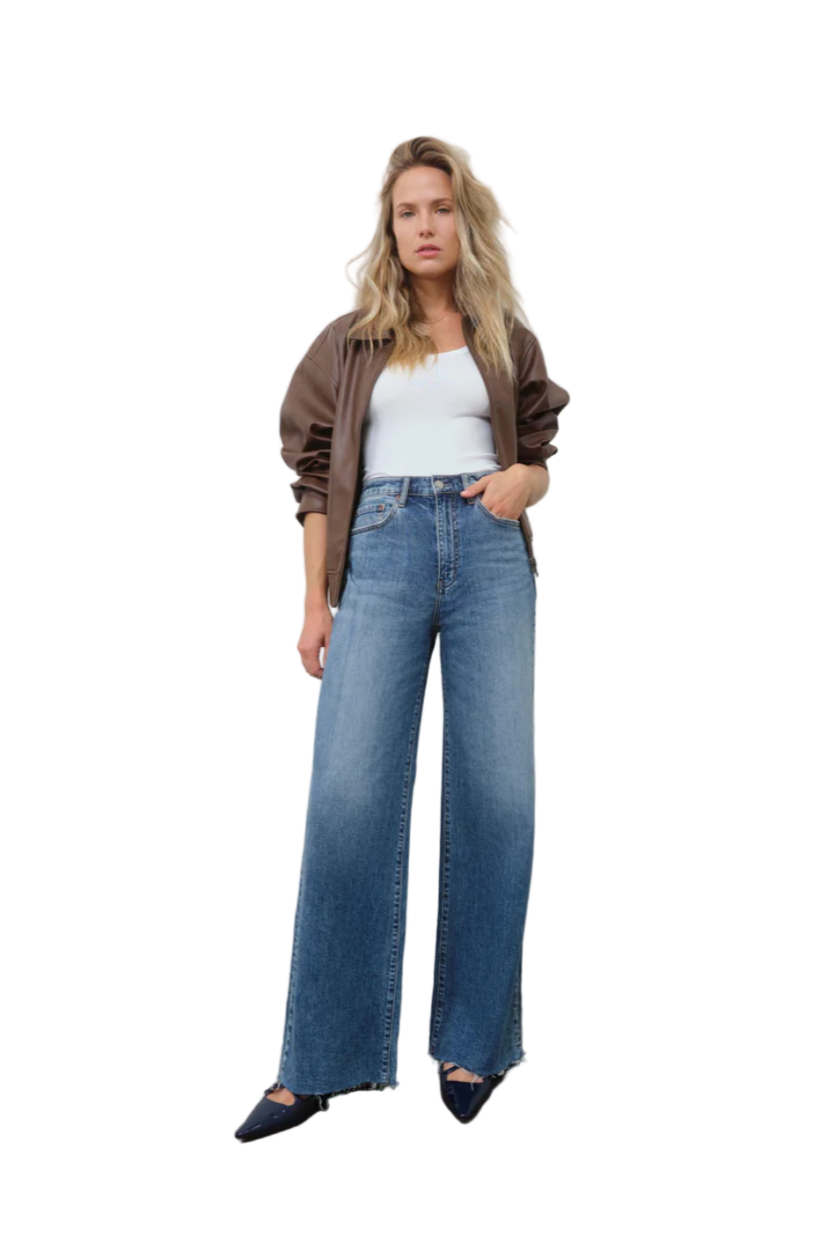 Woman wearing a white top, brown jacket, and blue jeans against a plain background