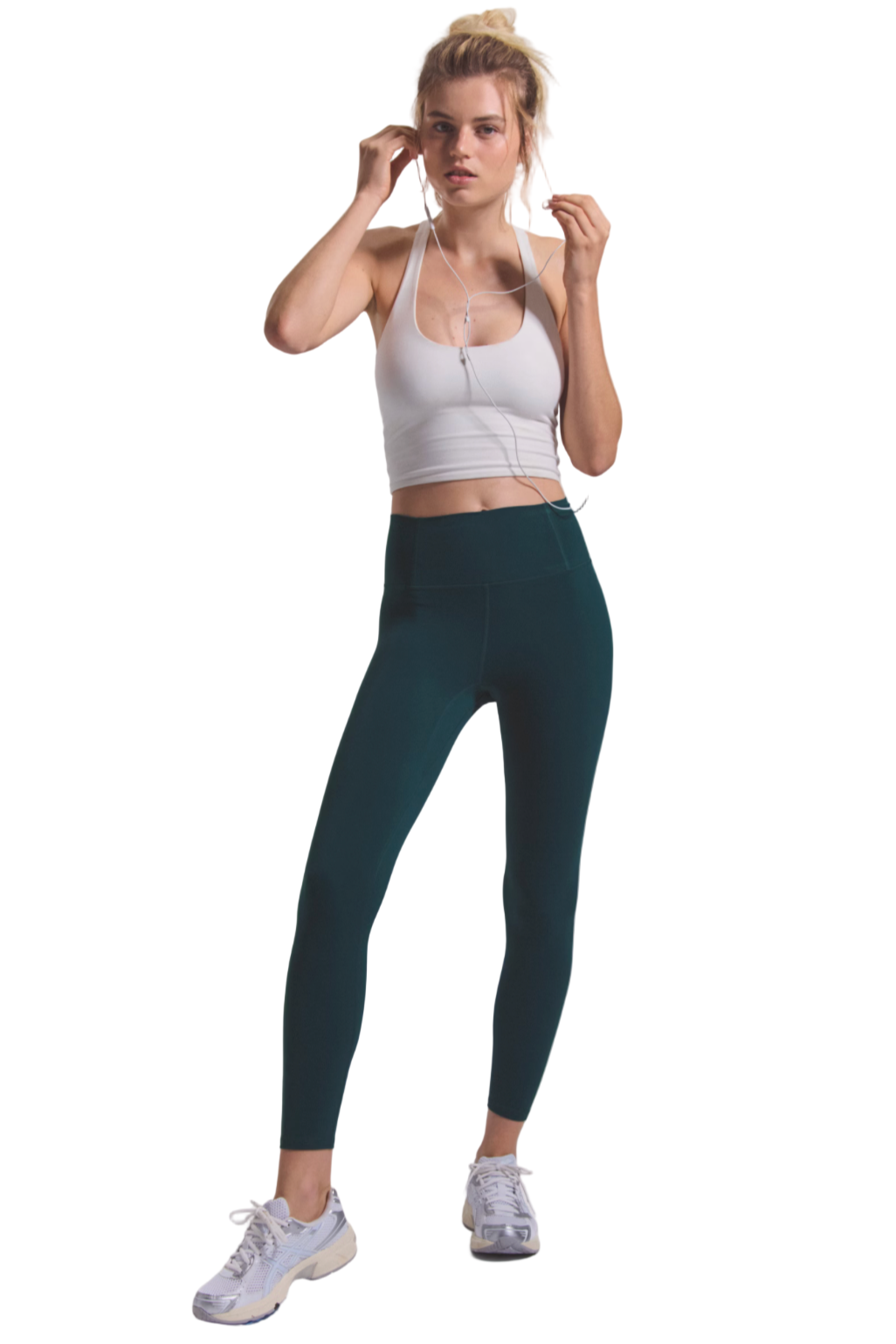 Woman wearing a white sports bra and dark green leggings on a plain background