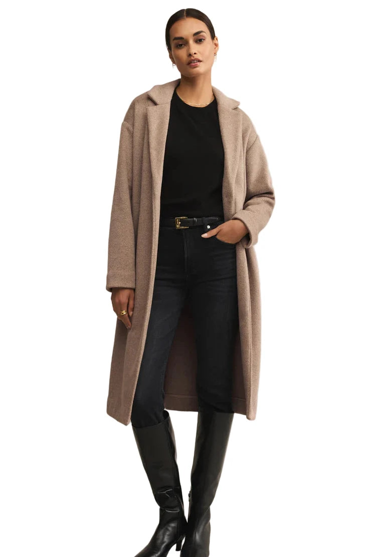Woman wearing a long beige coat, black top, jeans, and black boots on a beige background